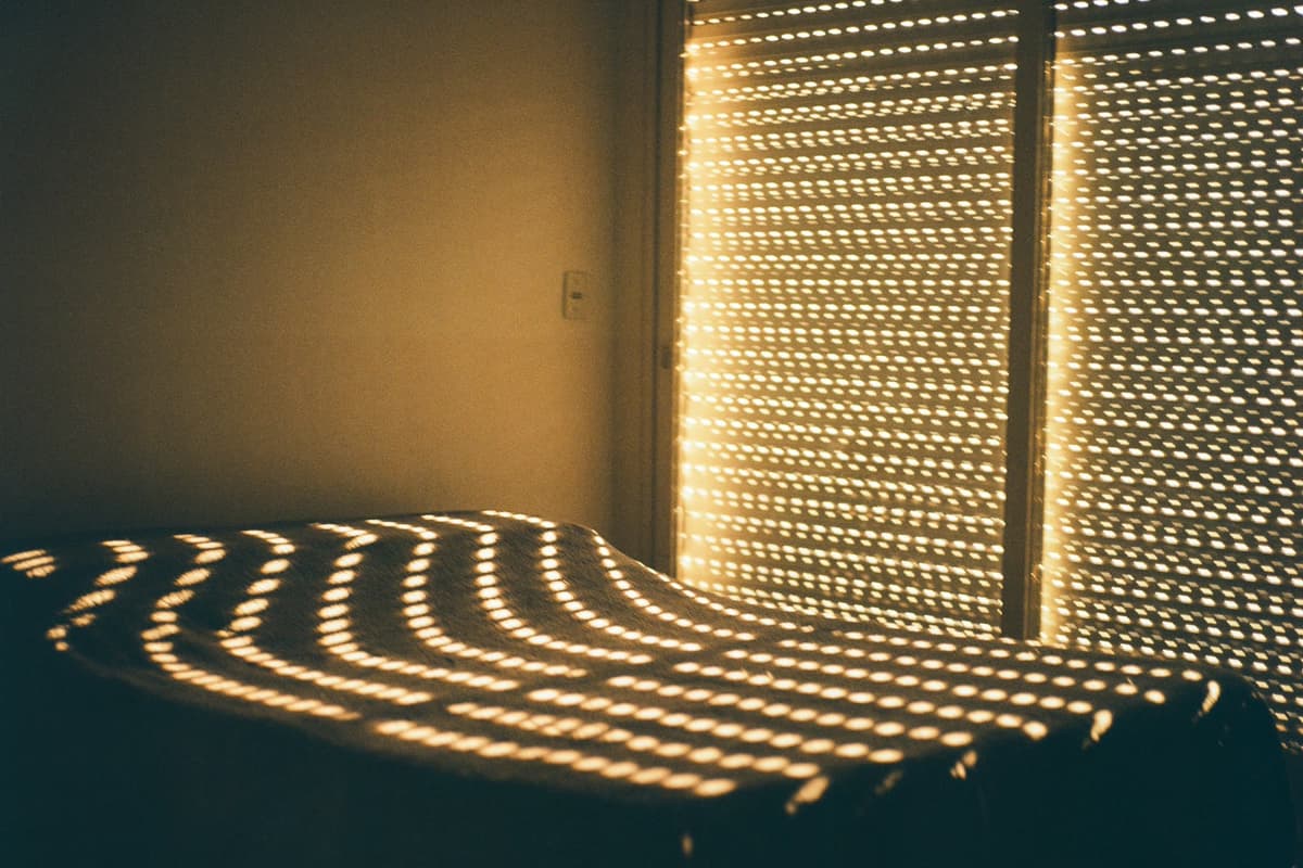 Warm sunlight through a perforated window shutter casting rows of light and shadow across a bed and wall